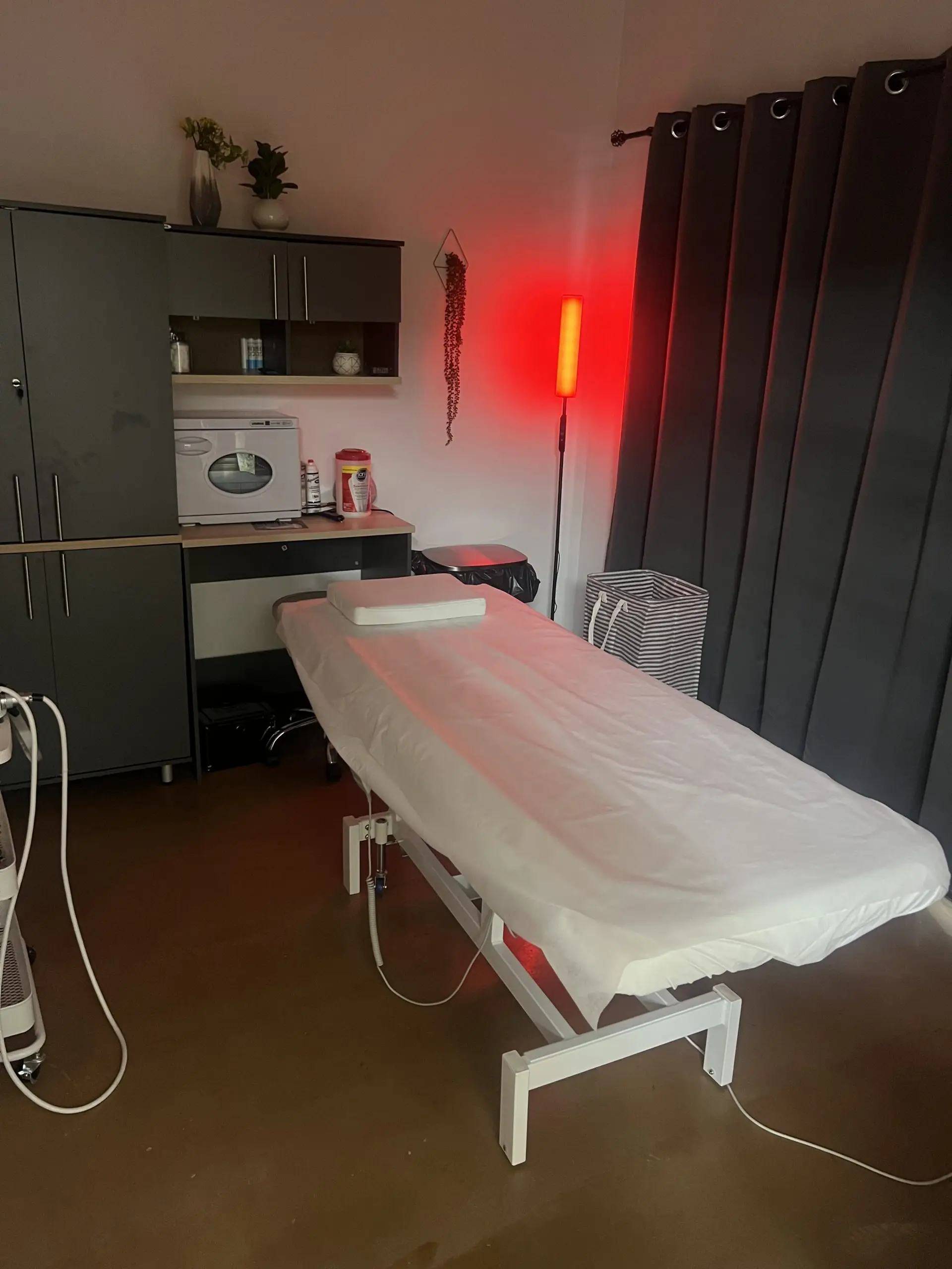 Private esthetic treatment suite with white treatment bed, modern storage cabinets, red lighting, and amenities.